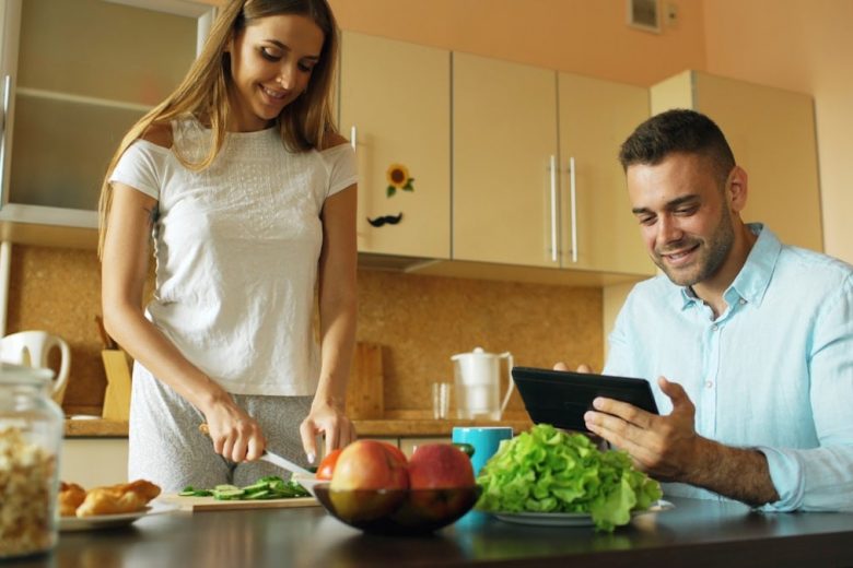 Attractive couple chatting in the kitchen early morning. Handsome man using tablet while his girlfriend cooking breakfast at home